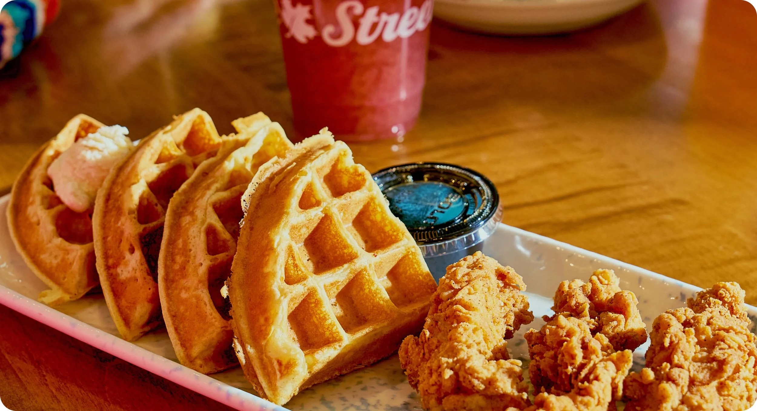 Chicken and waffles on a plate with a cup of syrup and a branded drink cup on a wooden table