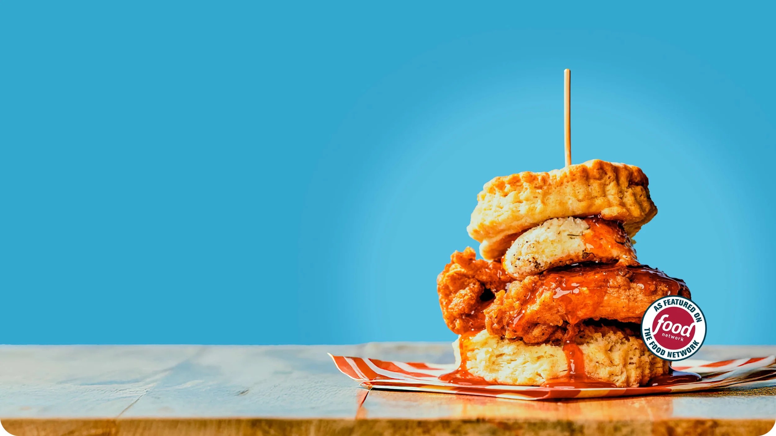 A tall fried-chicken biscuit with hot sauce dripping onto striped paper, on a bright blue backdrop, beside an “As featured on the Food Network” seal